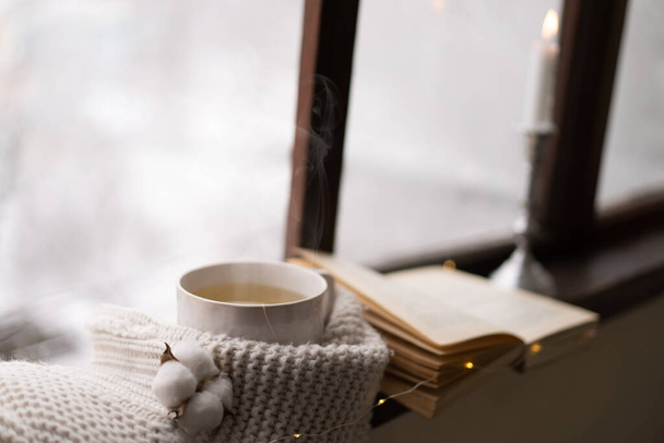 Cozy winter still life. Cup of hot tea and an open book with a warm sweater on a vintage wooden windowsill. Cozy home concept. Sweet home. - Photo, Image