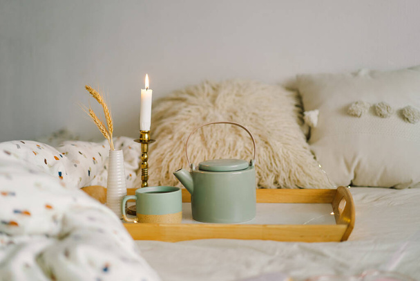 ベッドの上のサービストレイにお茶とティーポットのカップ。朝のベッドで朝食。自宅でお茶を楽しみ、自宅で時間を過ごすというコンセプト。居心地の良い秋や冬のコンセプト. - 写真・画像