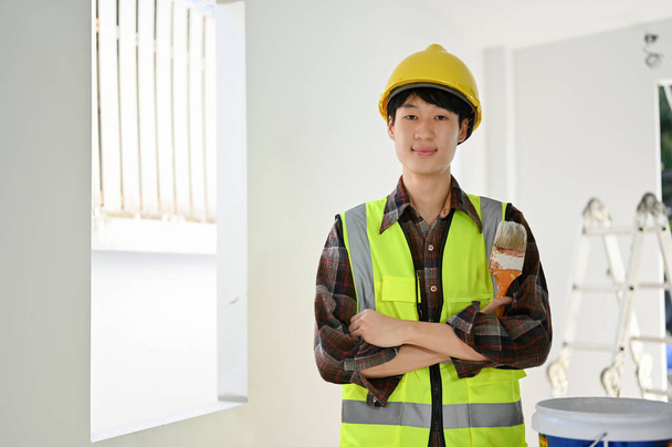 Розумний і професійний молодий азійський будівельник в уніформі і жовтих твердих стендах з перехрещеними руками, з пензлем фарби, посміхаючись і дивлячись на камеру. - Фото, зображення
