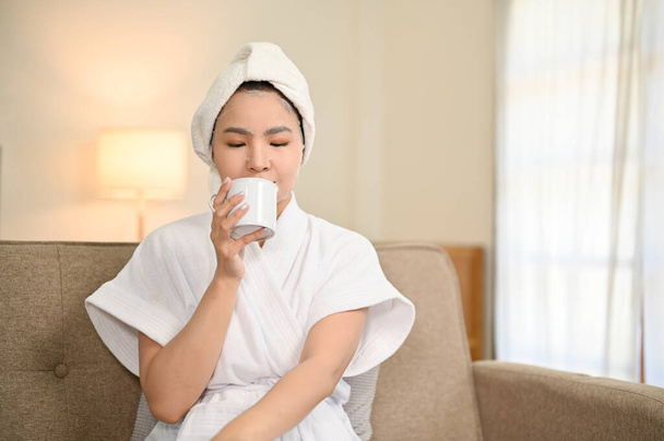 Расслабленная и расслабляющая молодая азиатка в халате потягивает утренний кофе на диване в своей гостиной. - Фото, изображение