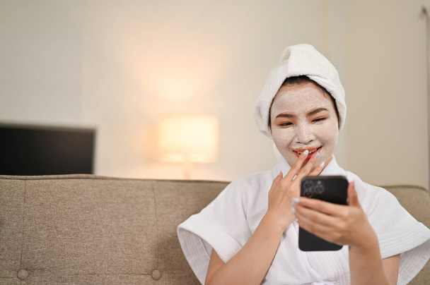 Joyful young Asian woman in bathrobe laughing on her phone, enjoys chatting with her friends while applying facial clay mask at home. - Photo, Image