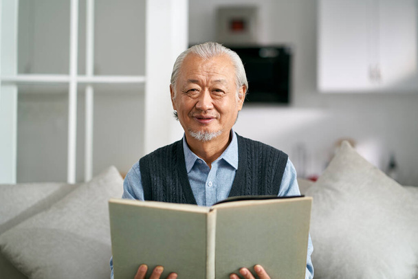 glücklicher asiatischer alter Mann sitzt auf der Couch zu Hause und liest ein Buch und schaut lächelnd in die Kamera - Foto, Bild