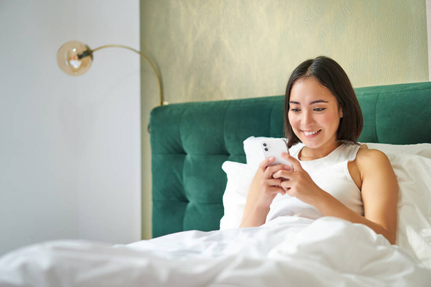 Krásná usměvavá asijská žena v posteli, čtení zprávy na smartphone a smích, těší líný den v ložnici, pomocí mobilního telefonu. - Fotografie, Obrázek