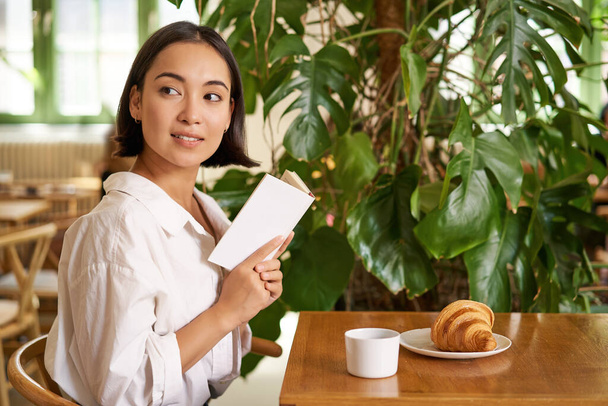 Piękna młoda Azjatka z książką w rękach, siedząca w kawiarni, pijąca kawę i jedząca rogaliki, uśmiechnięta, wyglądająca tajemniczo. - Zdjęcie, obraz