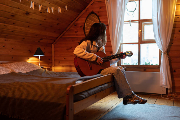 Melancholic lonely woman sits on bed with guitar in hands and looks out window admiring beautiful nature. Creative introverted girl is in cozy bedroom with wooden walls in attic of country house  - Fotoğraf, Görsel