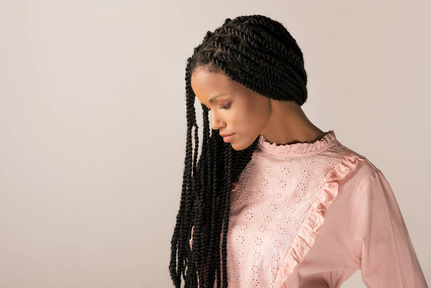 Joven hembra negra en blusa retro rosa con largas trenzas Afro mirando hacia abajo sobre fondo gris - Foto, imagen