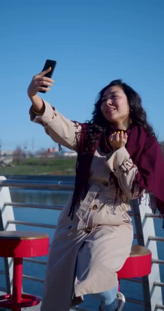 30 éves ázsiai nő egy online videóhívás során. A töltésen ül, és videokommunikáción keresztül kommunikál a beszélgetőpartnerrel. 4k felvétel. - Felvétel, videó