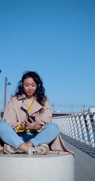Függőleges 4k felvétel egy fiatal ázsiai nőről meditál a töltésen. Fejhallgatóban ül, és pihentető zenét hallgat. Pihenj az irodai nyüzsgéstől és a városi zajtól. Városi életmód. - Felvétel, videó