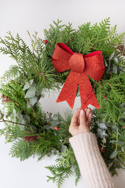 インテリアのためのスタイリッシュなクリスマスの花輪は、ドアに花輪を掛けます。手作りで、女の子は美しい赤い弓で新しく作られた花輪を持っています。祭りの雰囲気 - 写真・画像