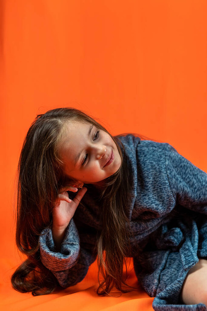 Pieni tyttö, jolla on tummat silmät harmaassa kaapussa, hymyilee istuessaan oranssilla taustalla. Eristetty. katsoo kehykseen. - Valokuva, kuva