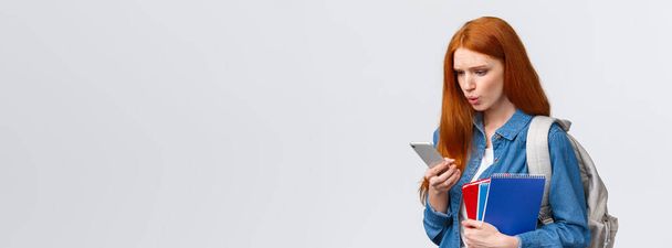What is that. Confused and displesaed. intense troubled redhead teenage, college girl with backpack and notebooks walking university and reading strange text on smartphone, white background. - Photo, Image