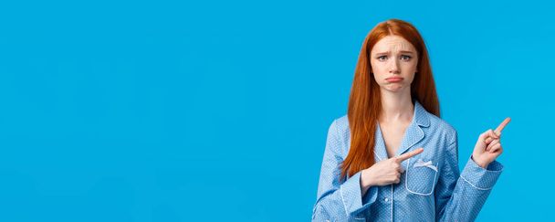 Regret, bad mood and advertising concept. Sad unhappy redhead girl in nightwear, sulking and frowning express disappointment or jealousy, pointing fingers right, standing blue background. - Photo, Image