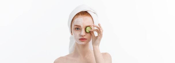 Aantrekkelijke jonge vrouw met mooie, schone huid. Wit masker en komkommers. Schoonheidsbehandelingen en cosmetologie spa therapie. Witte achtergrond. - Foto, afbeelding
