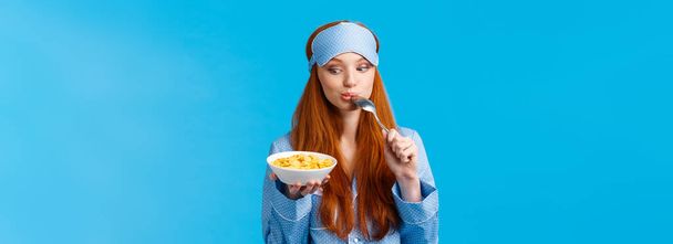 Τόσο νόστιμο. Χαριτωμένο και τρυφερό θηλυκό κορίτσι ξεκινώντας την ημέρα με υγιεινή διατροφή, τρώγοντας νόστιμα δημητριακά, γλείφοντας κουτάλι και κοιτάζοντας πιάτο δελεαστικό και πρόθυμο να το φάει, στέκεται μπλε φόντο. - Φωτογραφία, εικόνα
