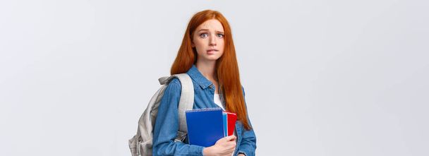 Worried cute and nervous redhead teenage girl scared parents scold her for skipping class, biting lip anxiously looking camera, holding notebooks and backpack over white background. - Photo, Image