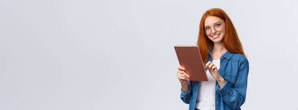 Taille-up portret vriendelijk gelukkig roodharige vrouwelijke student browsen online winkels, het houden van digitale tablet, het kiezen van nieuwe internet cursus voor het leren, glimlachen zorgeloos uitziende camera, witte achtergrond. - Foto, afbeelding