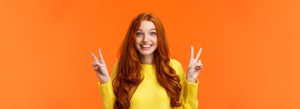 Paist-up portret piękny i wesoły ładna ruda kobieta pokazując gesty spokoju, uśmiechnięty aparat fotograficzny, podróż za granicę podczas ferii zimowych pozowanie w pobliżu zwiedzania, pomarańczowe tło. - Zdjęcie, obraz