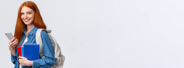 Charismatique rousse européenne à l'allure conviviale étudiante universitaire, collégienne avec sac à dos et cahiers, rentrant chez elle après les cours, texto ami sur smartphone, caméra sourire. - Photo, image