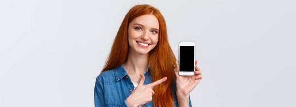 A lány ezen az alkalmazáson találta meg a randiját. Vidám vonzó vörös hajú nő kék szemekkel tanácsot ad, mutatja okostelefon kijelző és mutató ujj mobil képernyő, mosolygós ajánlást adni. - Fotó, kép