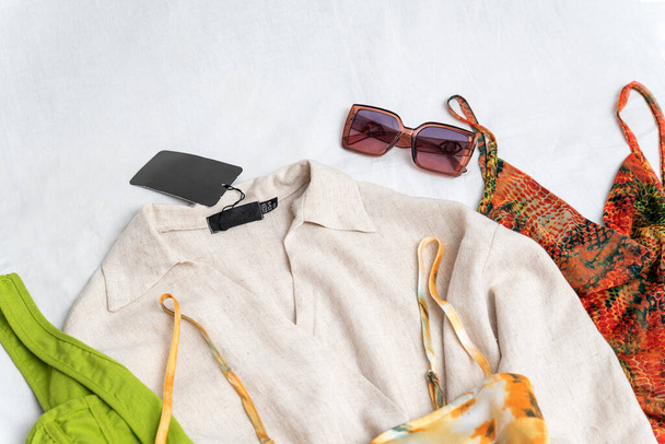 Robes colorées et chemise en coton ou lin avec étiquette noire vide. Armoire d'été lumineuse et lunettes de soleil sur fond blanc - Photo, image