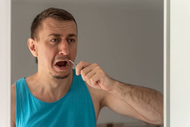 Portret van de mens in blauw T-shirt verwijderen van ongewenste haren in de neus staan voor de spiegel. - Foto, afbeelding