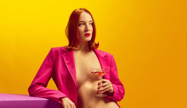 Drink aesthetics. Young redhead woman in pink jacket holding glass with champagne over yellow background. Concept of holiday, party, drink. Complementary colors. Copy space for ad. Pop art - Foto, afbeelding