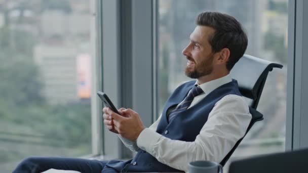 Satisfied entrepreneur watching phone receiving sms sitting at office in elegant suit close up. Confident bearded business man reading good news on smartphone screen. Manager taking break from work. - Footage, Video