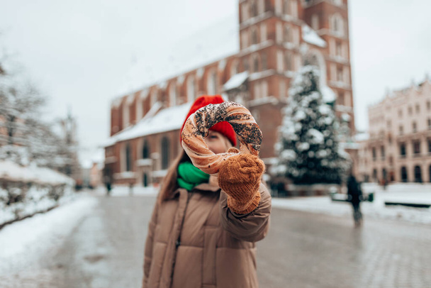 Vánoční turistka v jasném klobouku a teplém saku drží pečené obwarzanek tradiční polské kuchyně snack bagel na Staroměstském náměstí na Krakově. Veselé Vánoce a veselé svátky.  - Fotografie, Obrázek