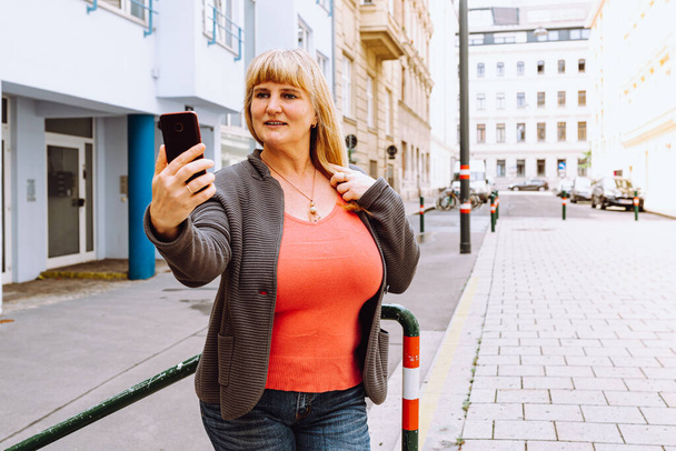 Middle-aged blonde busty woman takes selfie on smartphone camera, standing near house, against blurred background of residential modern area. Portrait of attractive middle age woman taking selfie in city - Photo, Image