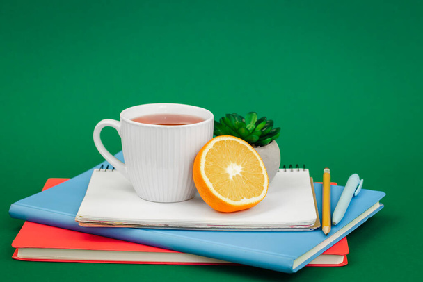 Cuadernos, una taza de té y una naranja sobre un fondo verde, el concepto de una merienda escolar. - Foto, Imagen