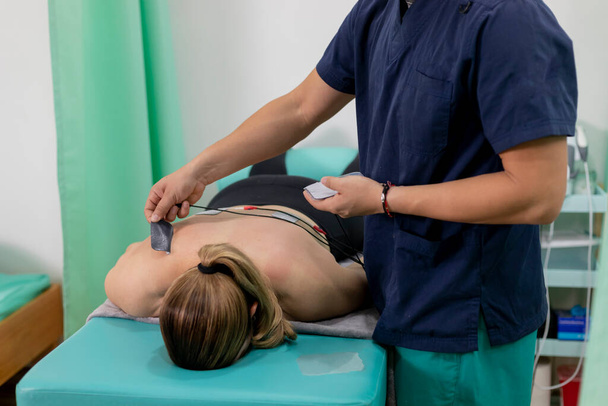 φυσικοθεραπευτής τοποθέτηση μαξιλαριών ηλεκτροδίων στην πλάτη μιας γυναίκας που είναι ξαπλωμένη σε φορείο - Φωτογραφία, εικόνα