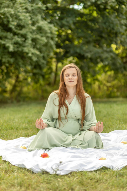 Белая беременная женщина медитирует в парке, сидит на одеяле, снимает стресс, внимательность. Наслаждаюсь лугом, расслабляюсь. Здоровый образ жизни. Труд, самозабота. Свежий воздух, зеленые деревья на заднем плане. - Фото, изображение
