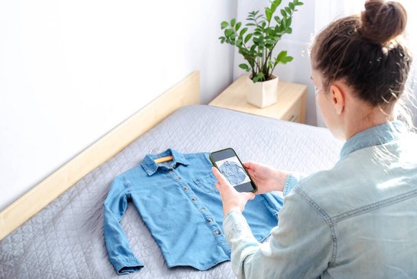 Une femme prend une photo sur smartphone de ses propres vêtements à vendre en ligne à la maison. Des vêtements usagés. Mode écologique et durable. Photo de haute qualité - Photo, image