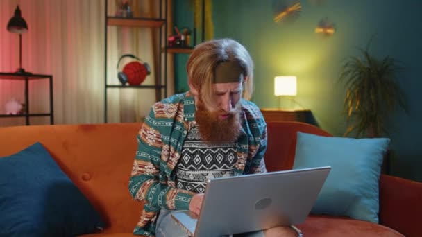 Displeased hippie bearded man use laptop typing browsing working, loses becoming surprised sudden lottery results bad news, fortune loss fail. Young hipster ginger guy at home in evening room on couch - Footage, Video