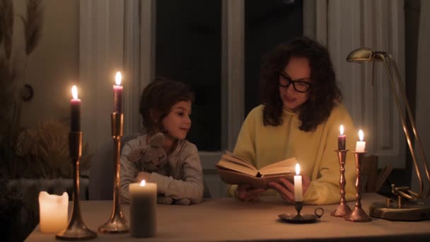 mère lit un conte de fées à sa fille à la maison aux chandelles dans un appartement sombre sans électricité en raison de l'agression militaire - Séquence, vidéo