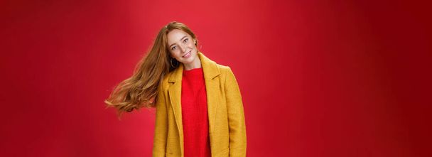 Очаровательная рыжая женщина с веснушками, машущими волосами. Улыбаясь широко наклоненной головой, как прическа летит в воздухе, представляя восторженные и беззаботные на красном фоне в теплом желтом пальто и трикотажное платье. - Фото, изображение