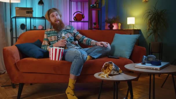 Aufgeregt sitzt der bärtige Hippie-Junge auf dem Sofa und isst Popcorn-Snacks und schaut sich zu Hause interessante Fernsehserien, Sportspiele, Filme und Social-Media-Filminhalte an. Kerl genießt häusliche Unterhaltung - Filmmaterial, Video