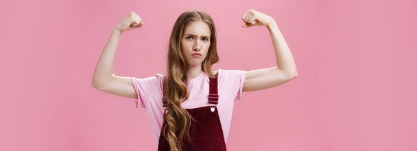 Chica muestra ella fuerte e independiente. Mujer delgada y segura de aspecto serio levantando las manos para mostrar los músculos sintiéndose segura de sí misma en su propio poder y las fortalezas femeninas posando contra la pared rosa. Copiar - Foto, Imagen