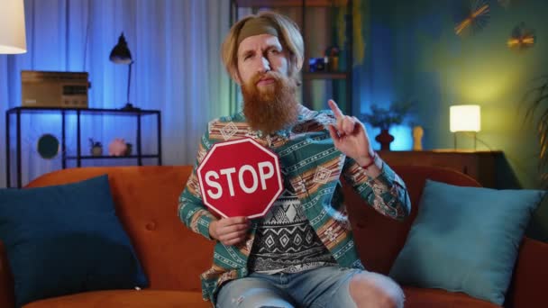 Bearded hippie redhead man at home couch say no hold showing stop inscription banner, warning of finish, prohibited access, declining communication, danger. Young hipster guy in night evening room - Footage, Video
