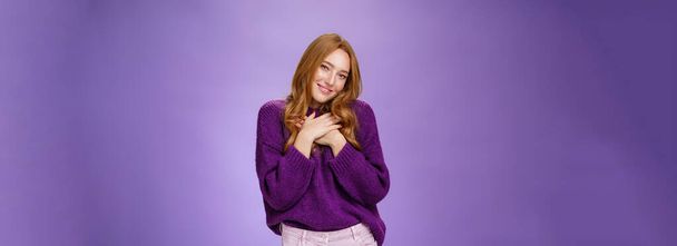 Qué lindo agradecimiento. Chica pelirroja encantadora tierna en suéter púrpura cogida de la mano en el corazón cabeza inclinada coqueta y feliz como sonriendo a la cámara agradecida y tocada con regalo romántico sobre la pared violeta. - Foto, Imagen