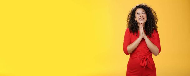 Lifestyle. Dreamy excited cute b-day girl with curly hair in cute red dress rubbing palms together near chest as hands in pray smiling looking up delighted and hopeful making wish over yellow - Photo, Image
