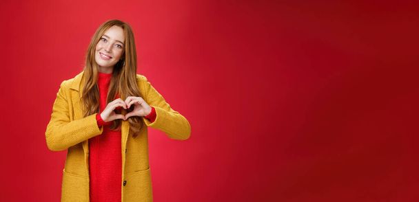 Любовь, романтика и осенняя концепция. Портрет очаровательной нежной и нежной молодой рыжей женщины в желтом пальто, показывающей сердечный жест, делающий признание в симпатии, улыбающейся мило на камеру через красную стену. - Фото, изображение
