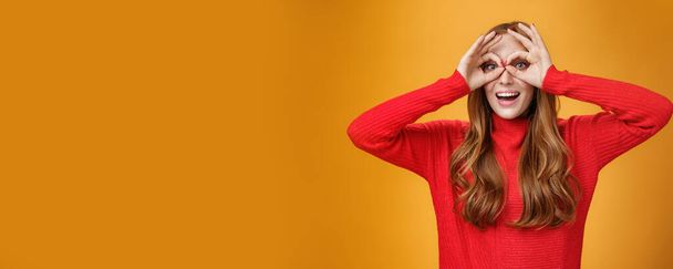Taillierte Aufnahme eines kindisch-verspielten, lustigen Ingwermädchens in rotem Strickkleid, das Gesichter macht, die Kreise über den Augen wie eine Brille zeigen und breit lächelnd Spaß vor orangefarbenem Hintergrund hat wie ein Kind - Foto, Bild