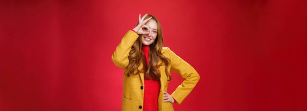 Helder en zonnig meisje voelt zich gelukkig, zelfs op regenachtige dagen staan in gele stijlvolle jas over rode achtergrond tonen oke gebaar over oog, brede glimlach op camera, hand in hand op taille. - Foto, afbeelding