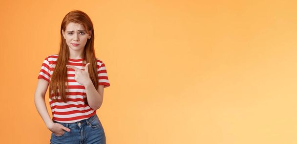 Upset uneasy cute ginger gitl long natural red hair, frowning smirking hesitant, complain, looking suspicious and doubtful point left, not sure if product really good, feel unsure, orange background. - Photo, Image