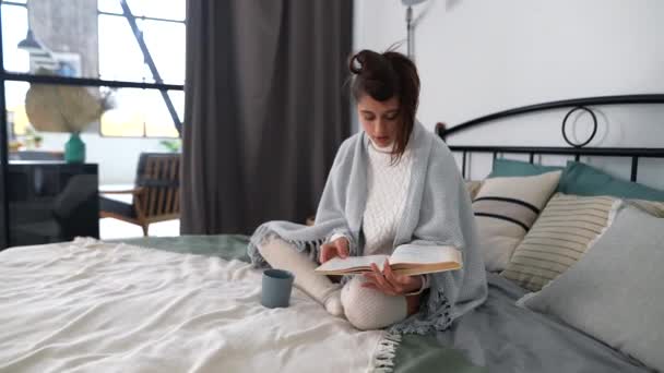 Μια νεαρή γυναίκα διαβάζει ένα βιβλίο ενώ κάθεται στο κρεβάτι στην κρεβατοκάμαρα. - Πλάνα, βίντεο