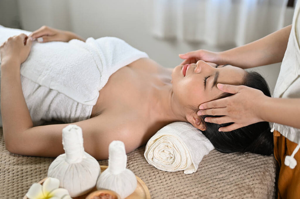 Mooie en ontspannen jonge Aziatische vrouw liggend op massagetafel, het ontvangen van Thaise gezichtsmassage door een professionele masseur in Thaise spa salon. - Foto, afbeelding