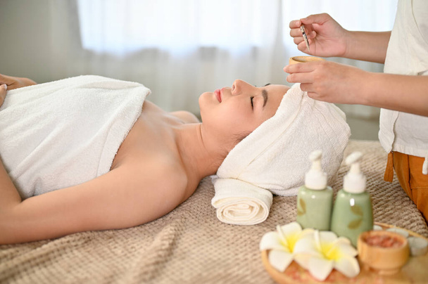 Mulher asiática bonita deitada na mesa de massagem, recebendo máscara de barro de tratamento facial por um trabalhador de spa. vista lateral - Foto, Imagem