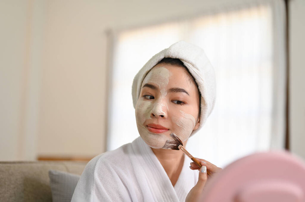 Очаровательная молодая азиатка в халате, смотрящая в зеркало, надевая детоксикационную маску на лицо дома. стиль жизни  - Фото, изображение