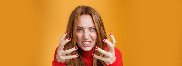 Close-up sot of pissed and angry redhead woman raising hands and clenching them with anger and outrage being irriated and annoyed grimacing from dislike, hate and pressure over orange wall. - Фото, изображение